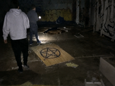 I thought the shape beneath the rotting wrestling mat was a pile of clothes until I saw the hand. Then I noticed the candles arranged around it, the copper-colored marks across the concrete, and a black symbol drawn over one bare ankle. We ran before any of us understood what we were looking at. Hours later, a detective would realize the strangest thing about that room was not what had been left there. It was how carefully someone had tried to make police believe the killing happened there.